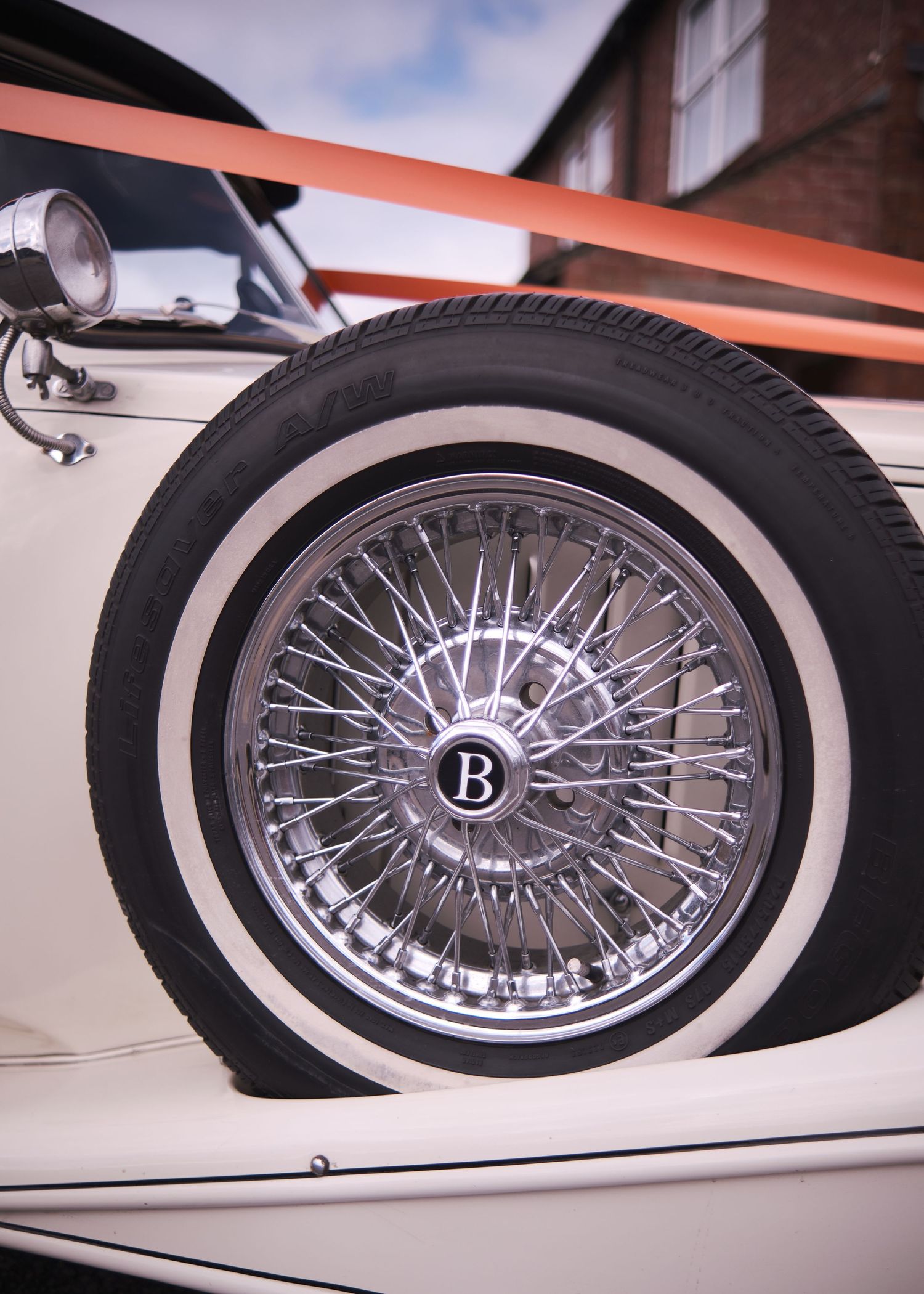 Close-up of a classic Bentley car wheel with chrome wire spokes and white wall tire parked outside a brick building.