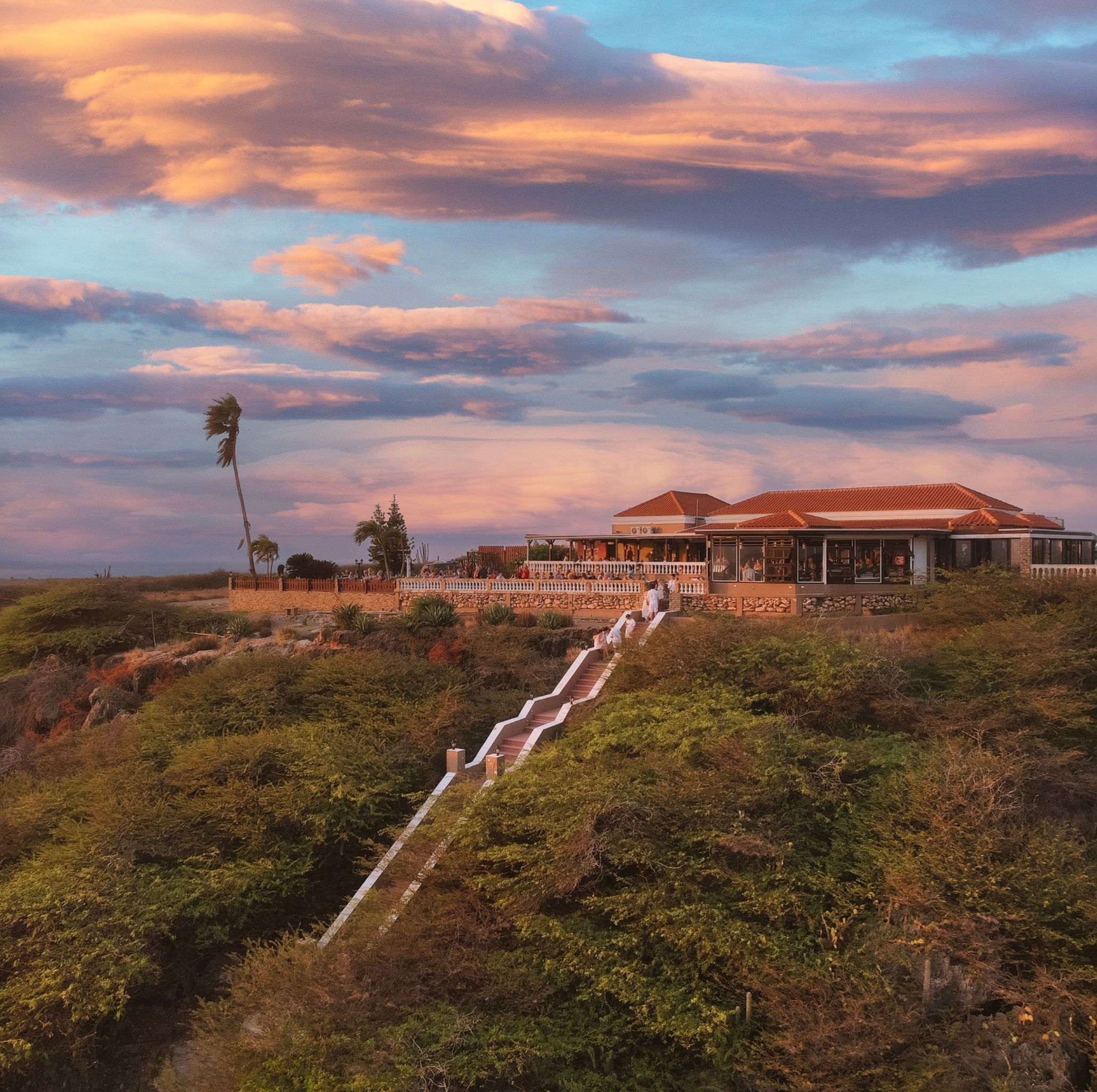 Palm trees and thatched beach cabanas illuminate the shoreline at sunset with dramatic purple and blue clouds over turquoise waters.