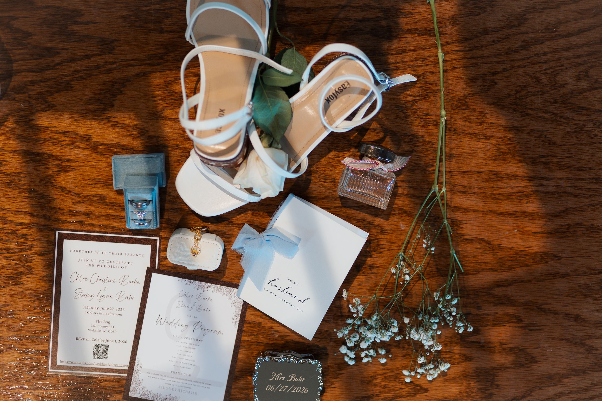 Wedding flat lay featuring white heels, invitations, perfume, ring box, and dried florals on a wooden surface.