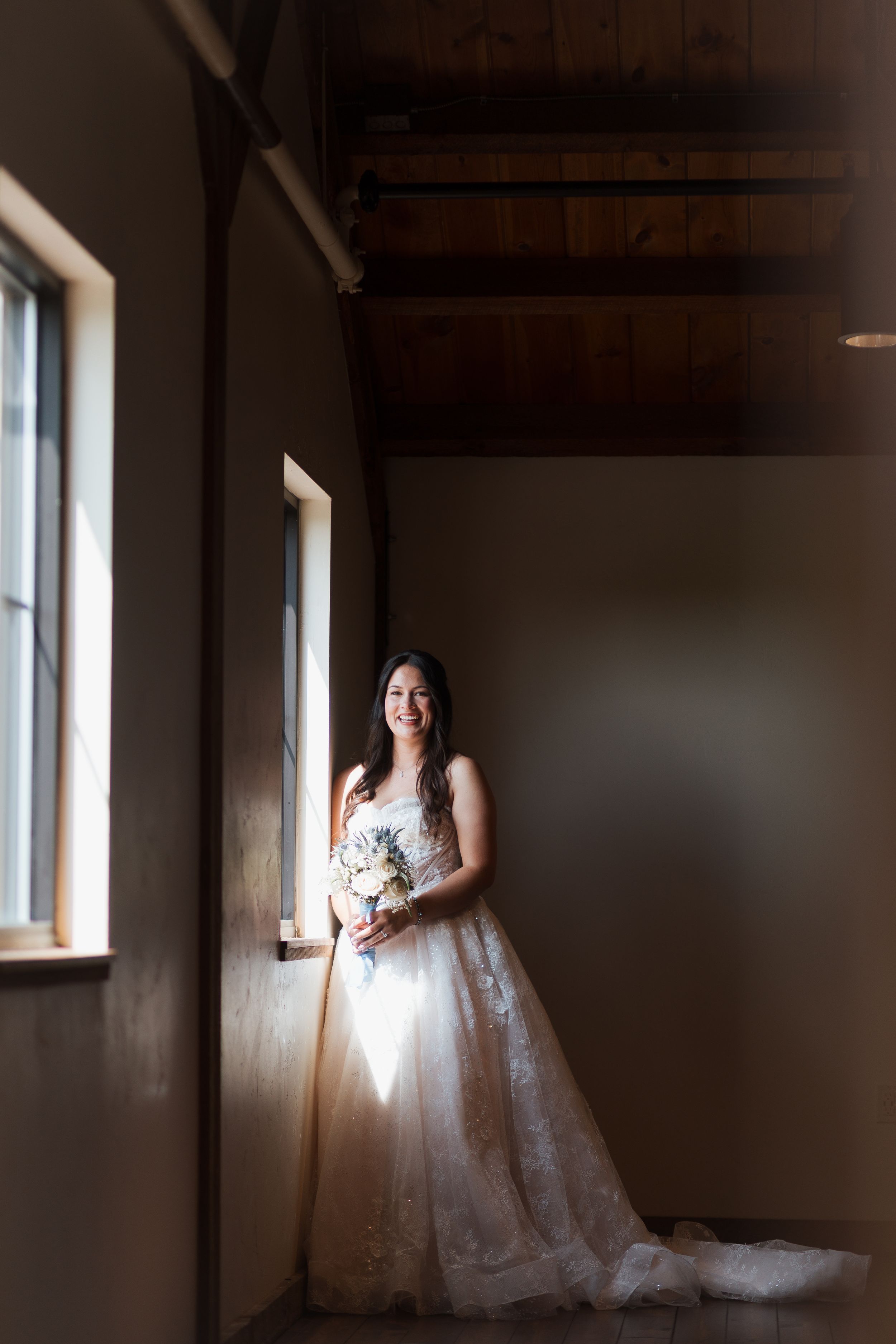 Bride in lace ball gown holding bouquet stands by sunlit window in rustic venue with wood ceiling.