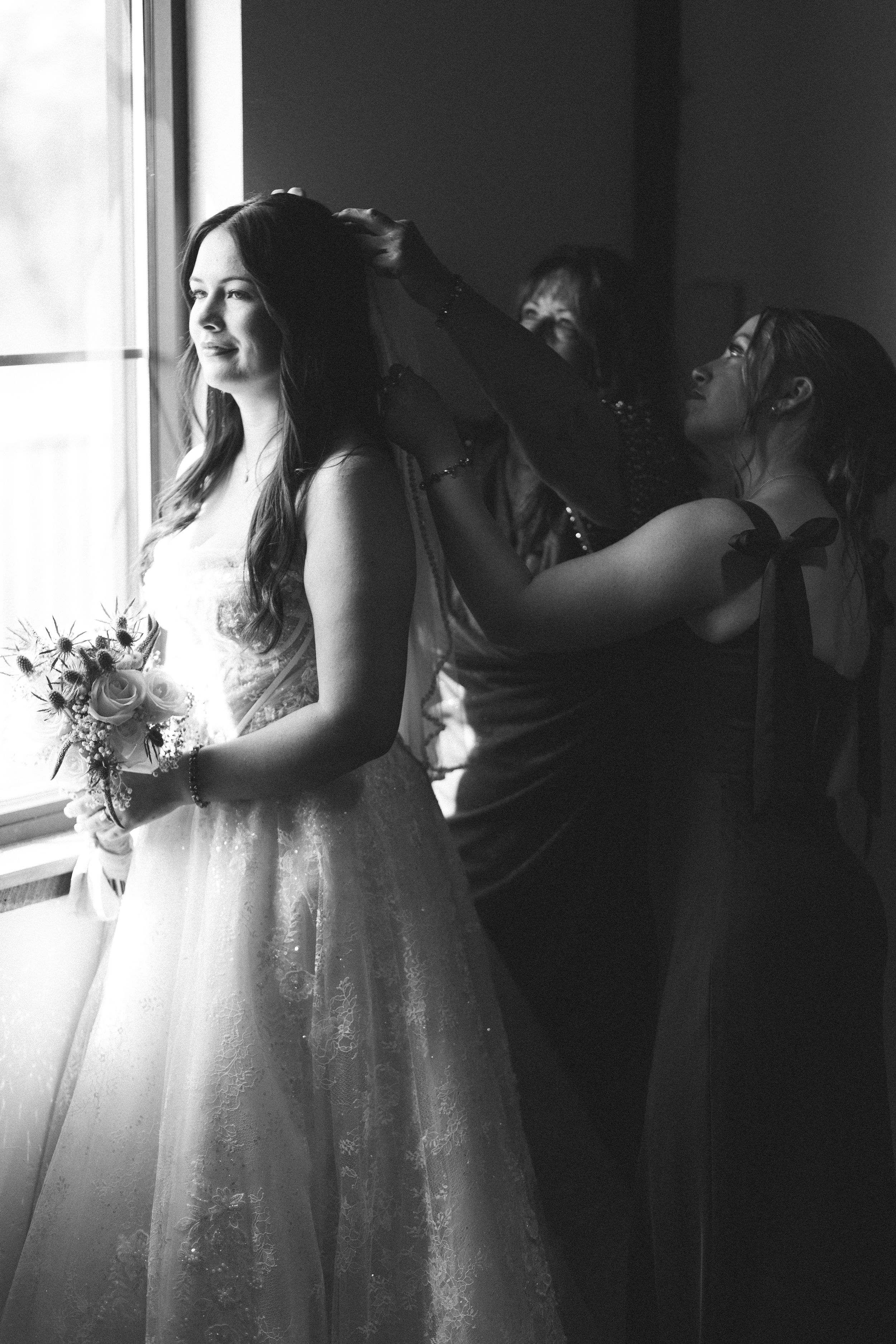 Black and white photo of a bride in a wedding gown holding a bouquet while bridesmaids help style her hair.