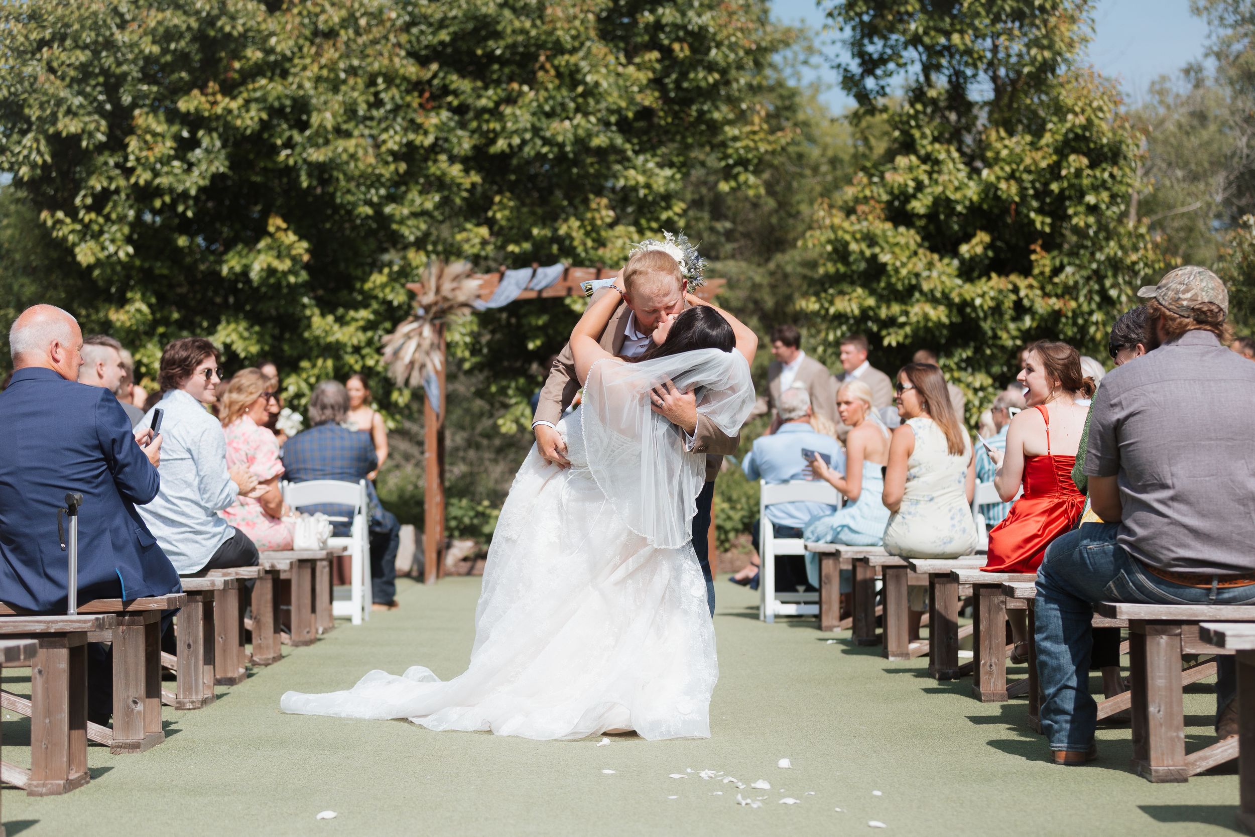Groom dips bride in a romantic kiss at outdoor wedding ceremony with guests seated on wooden benches.