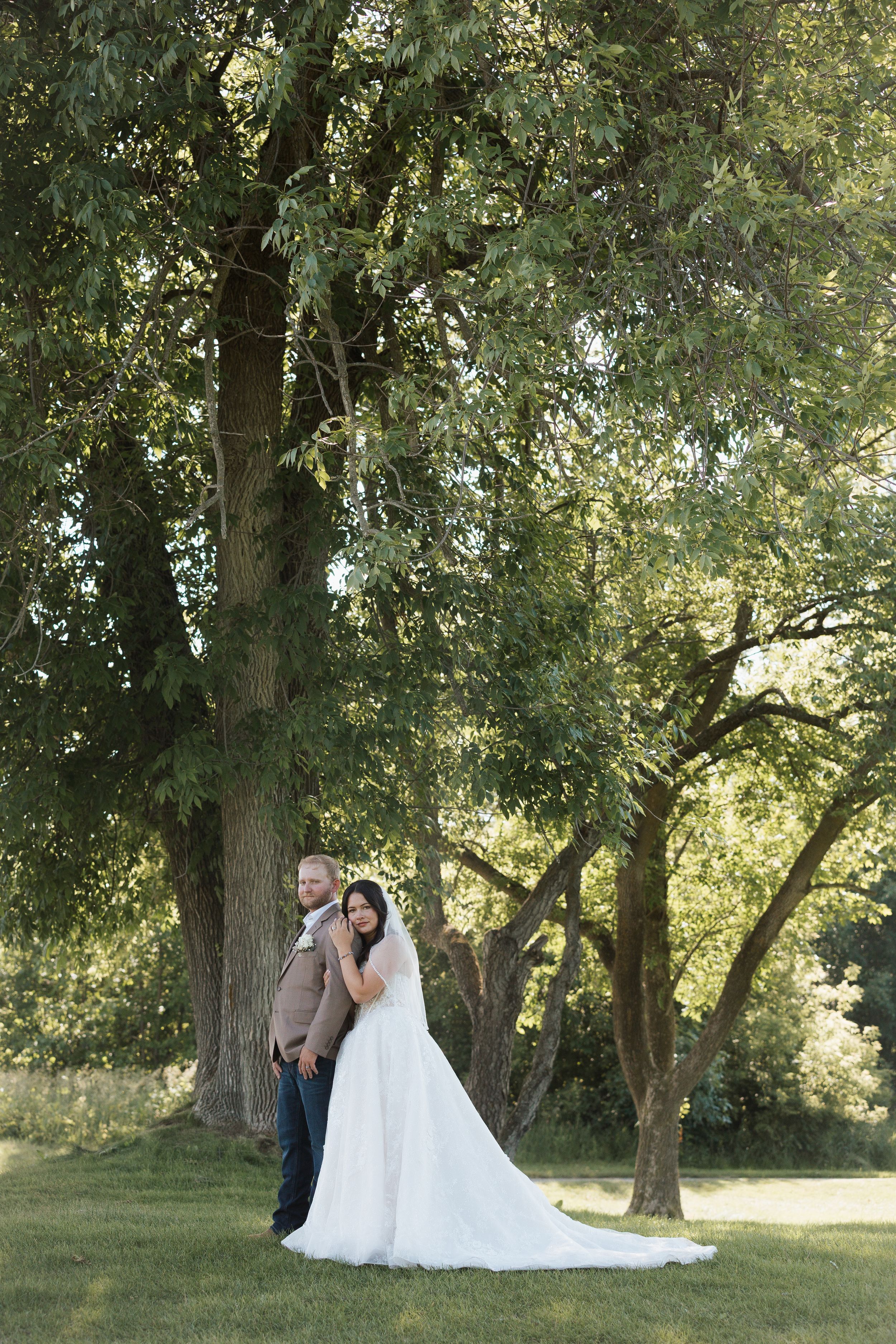 Bride and groom pose together under large green trees in a lush outdoor setting, bride wearing a flowing white gown.