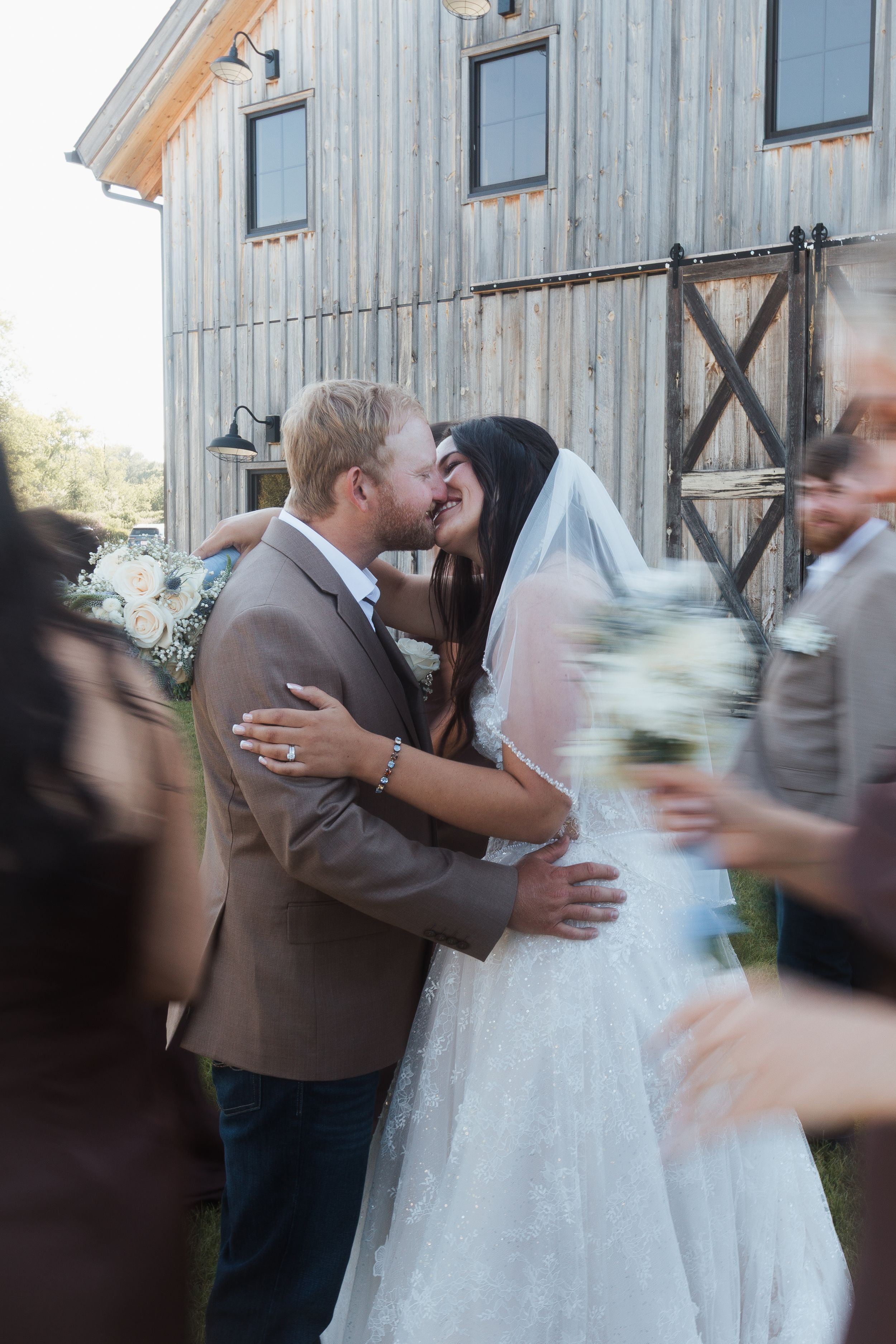 Bride and groom share first kiss at rustic barn wedding venue, bride in lace gown with veil holding white bouquet.