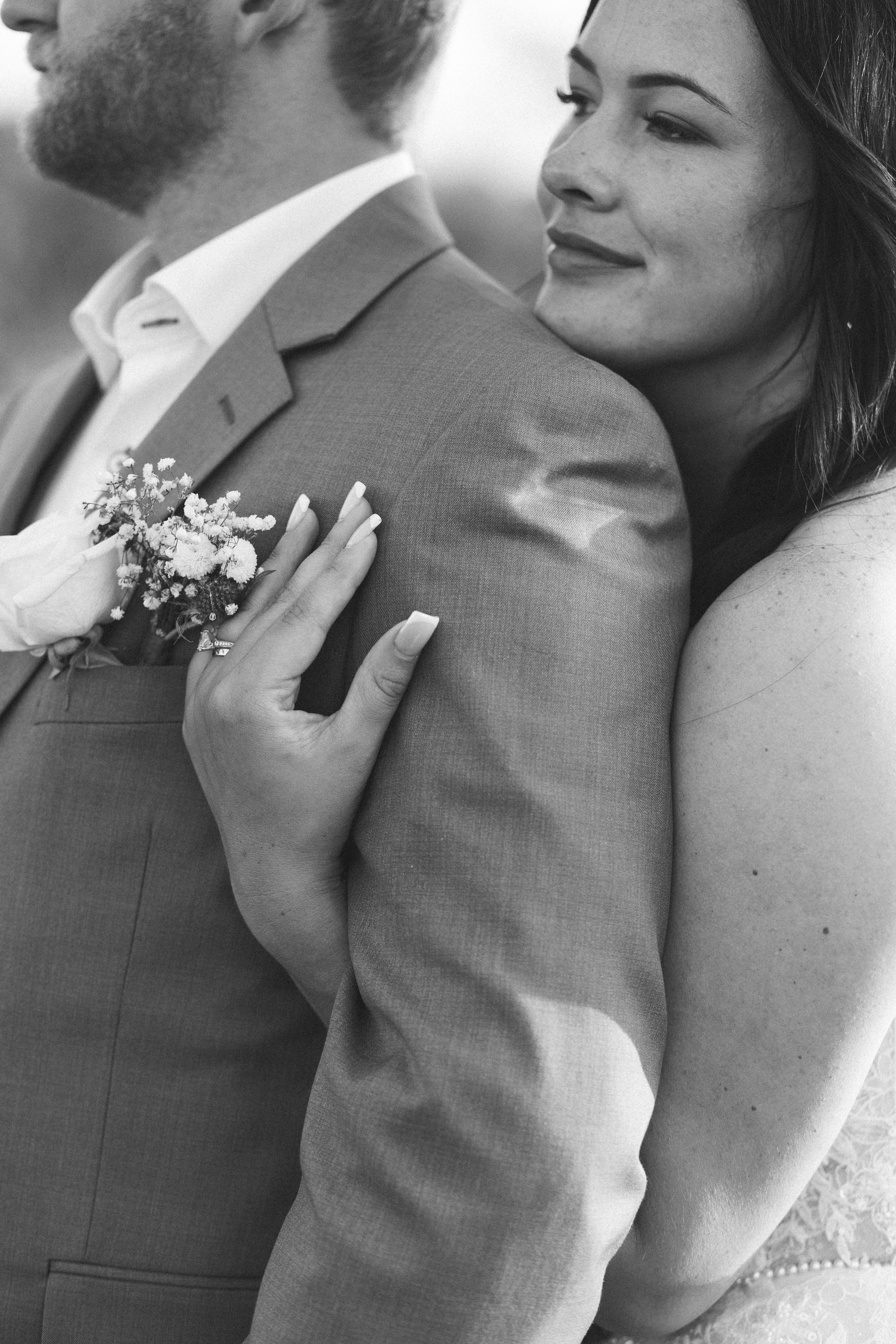 Black and white photo of a bride embracing groom in suit with boutonniere at wedding.