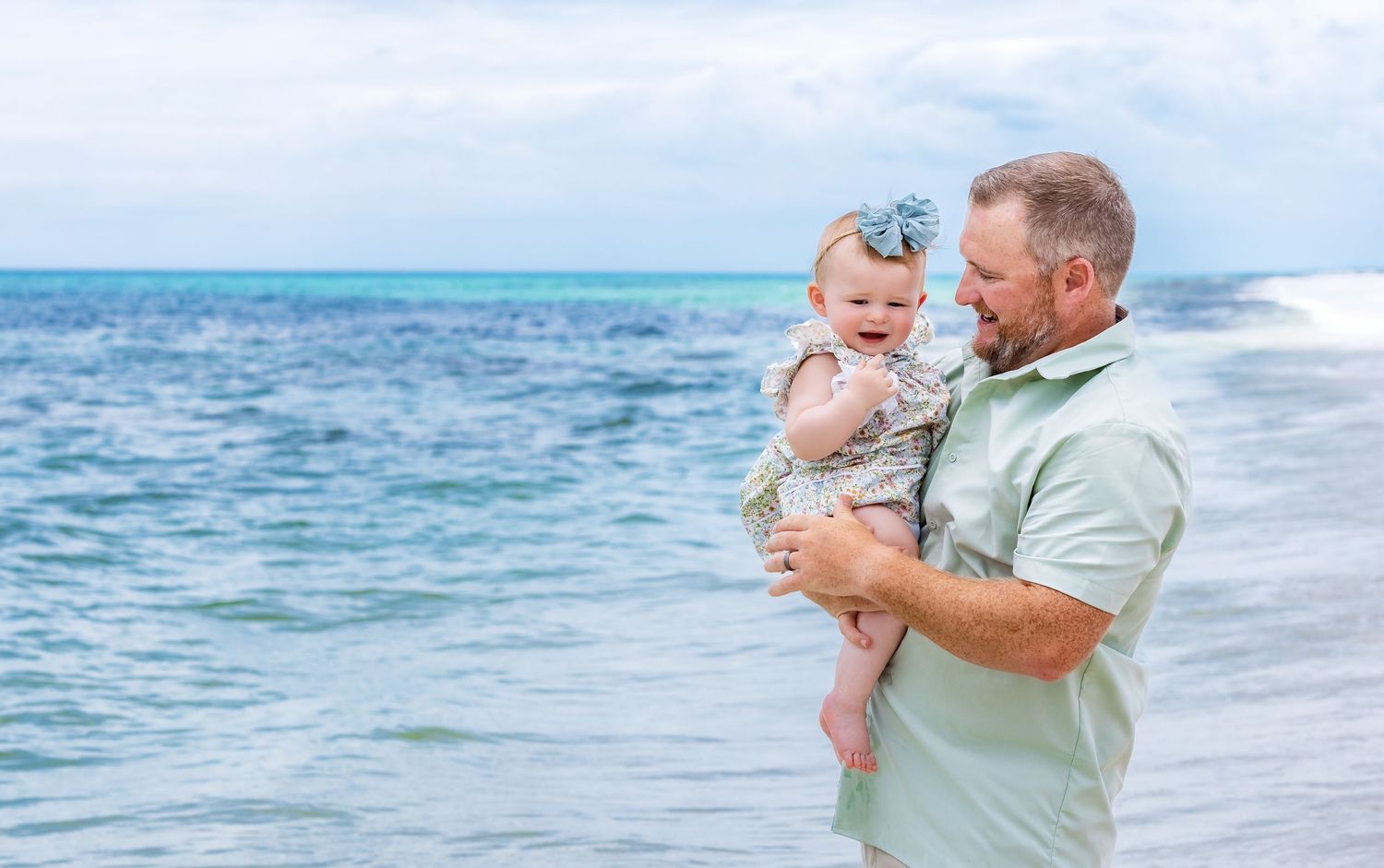 A man holding a laughing baby girl on a sunny beach with turquoise ocean waves in the background.