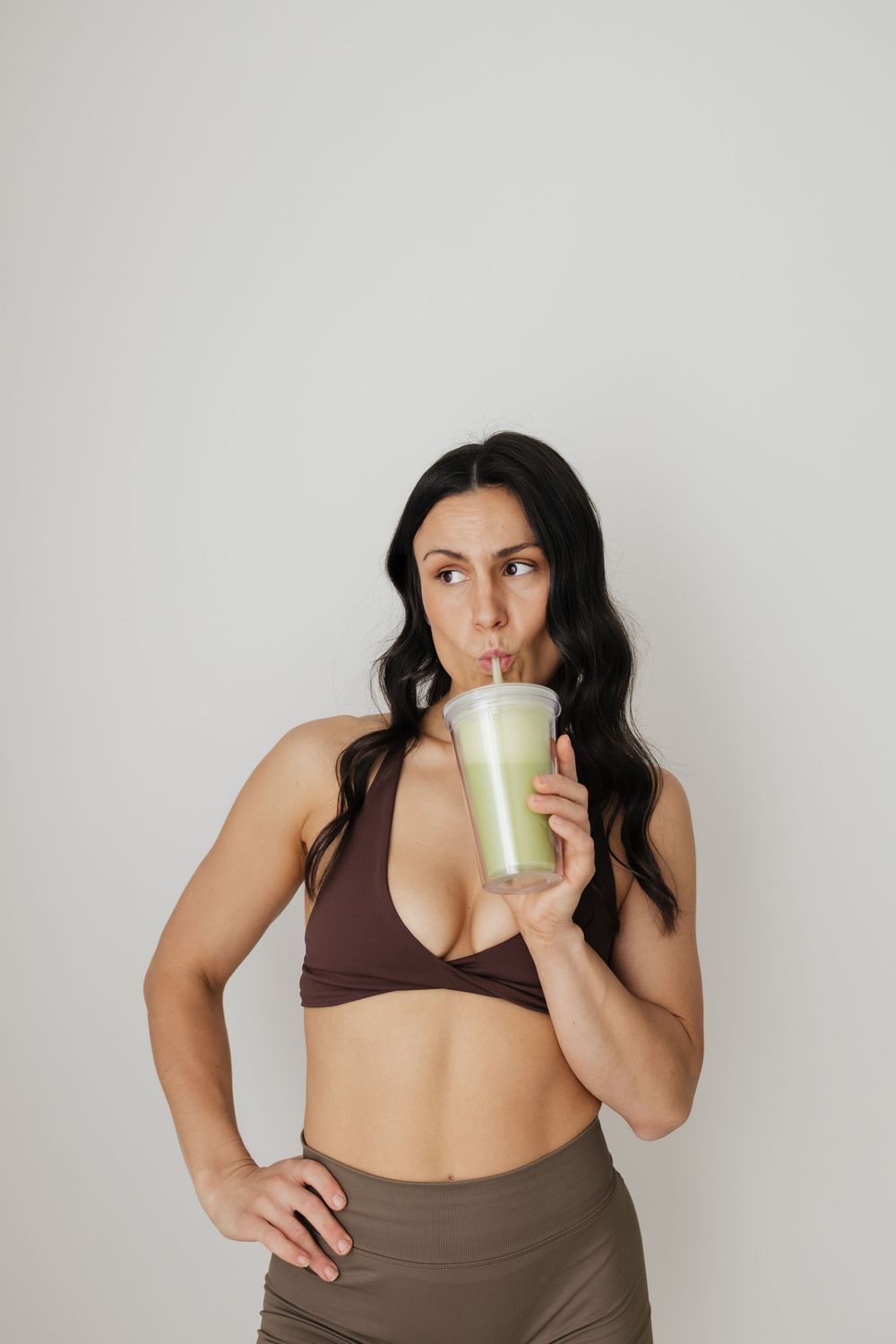 Woman in brown sports bra sipping a green smoothie, posing against a white background.