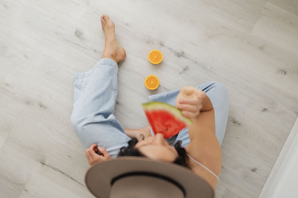 Woman sitting on floor eating watermelon slice with halved oranges nearby, wearing jeans and a hat.