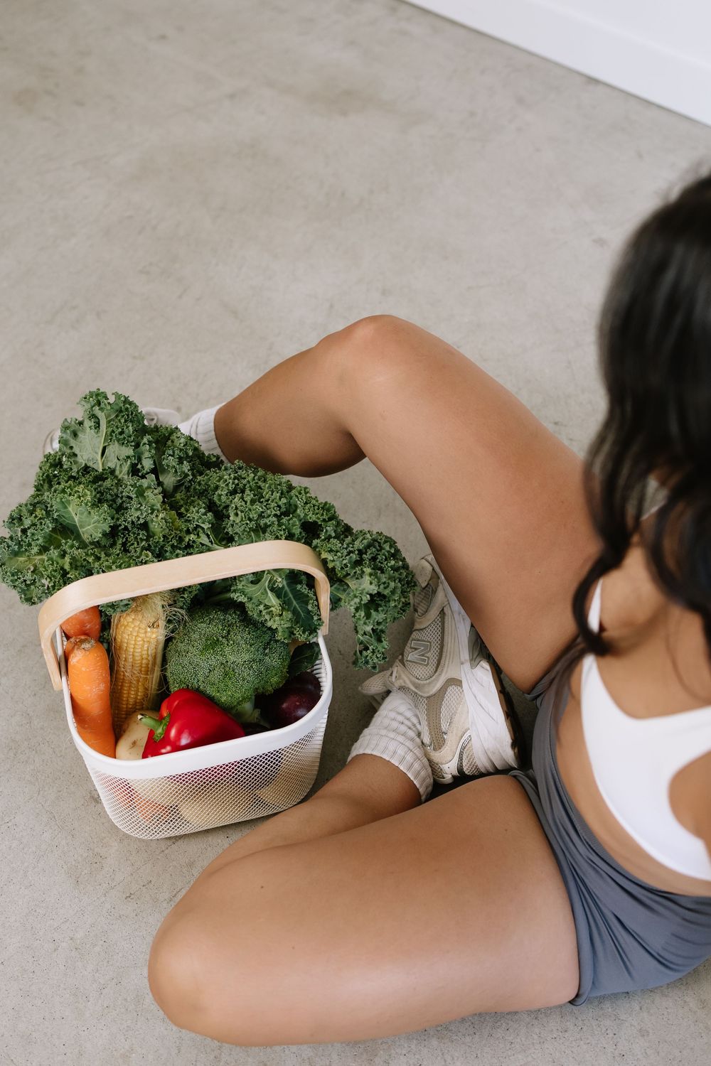 Woman sitting on floor holding a basket filled with fresh vegetables including kale, broccoli, corn, and peppers.