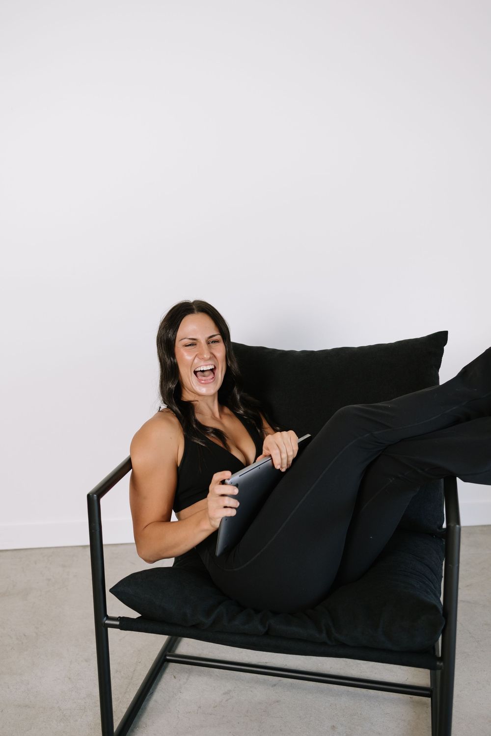 Woman in black outfit laughing while sitting in a modern black chair holding a tablet.