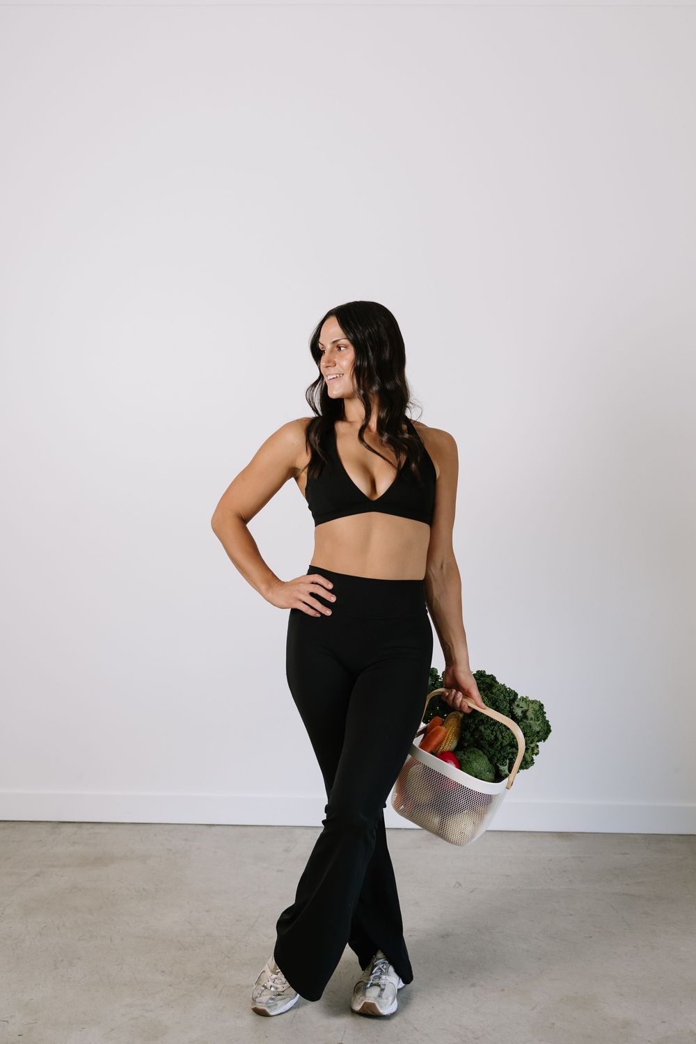 Fit woman in black crop top and flare pants holding a basket of fresh vegetables against white wall.
