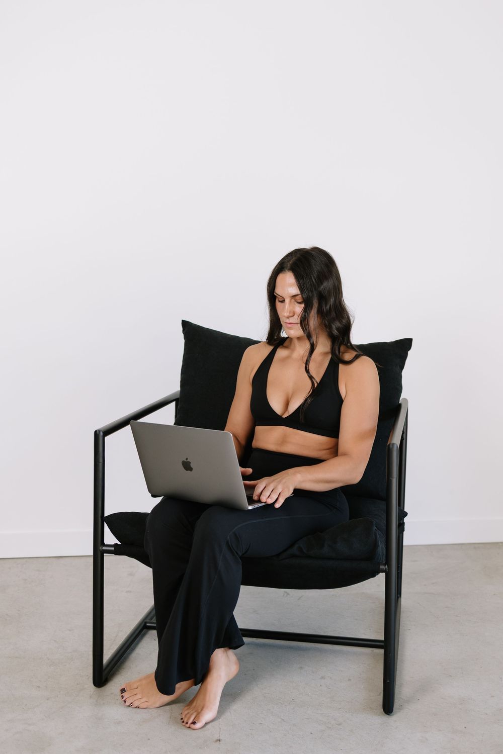 Woman in black athletic wear sits barefoot on modern chair, working on MacBook laptop against white wall.