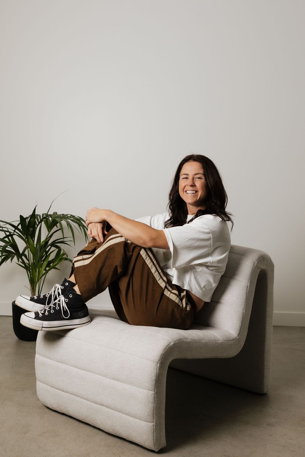 Woman smiling in white top and brown pants sitting relaxed in a modern cream armchair with a plant in the background.