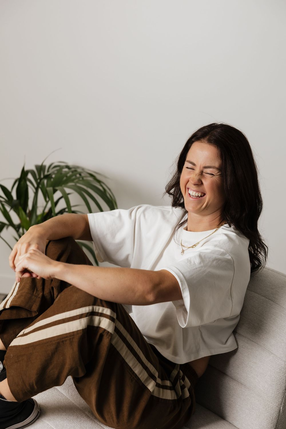 Woman laughing joyfully in white tee and brown striped pants, sitting relaxed on a couch near a green plant.