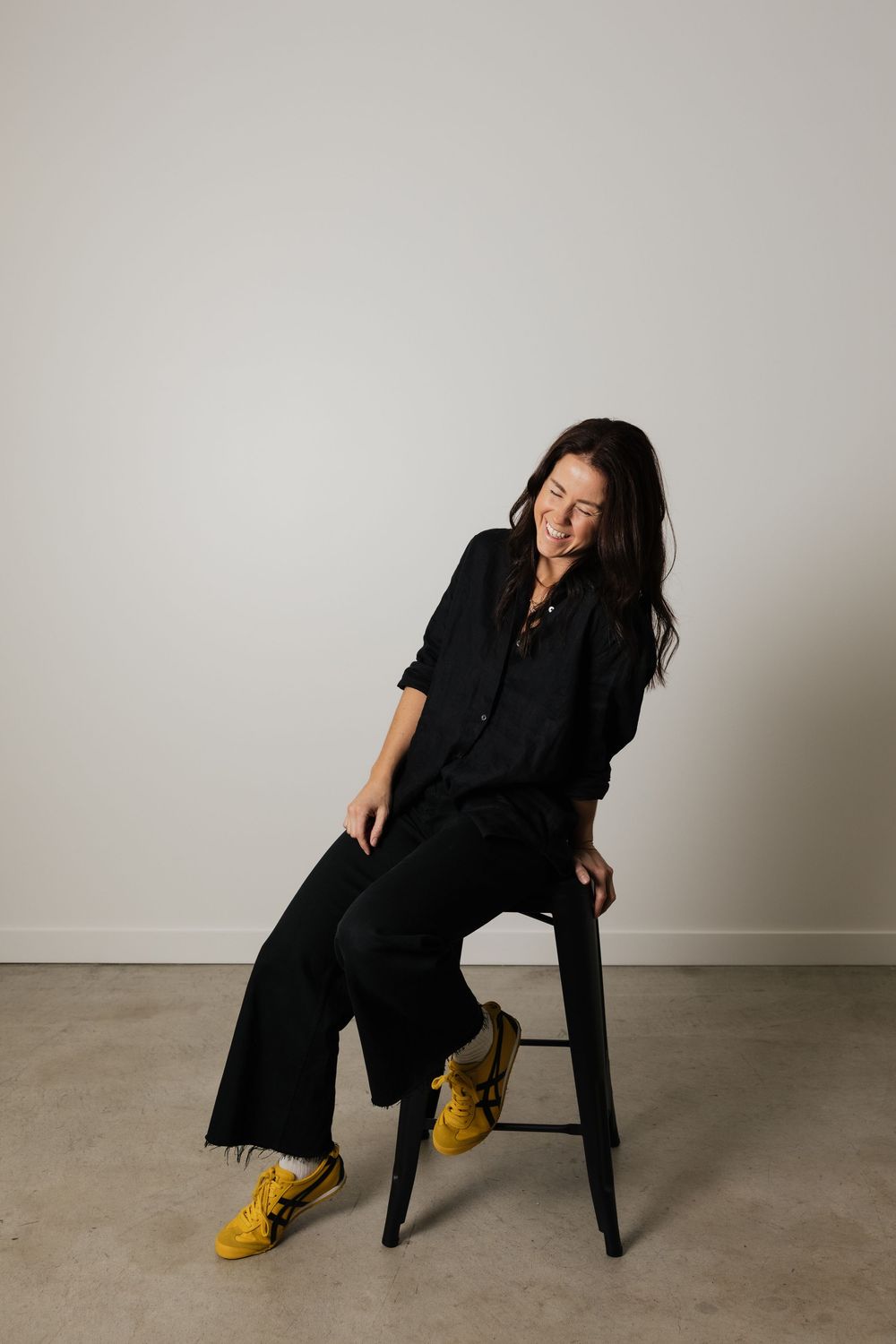 A smiling woman in all-black outfit with yellow sneakers sits on a stool against a light gray wall.