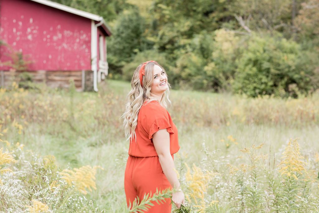 A young woman in an orange romper stands in a wildflower field near a red barn, smiling over her shoulder.