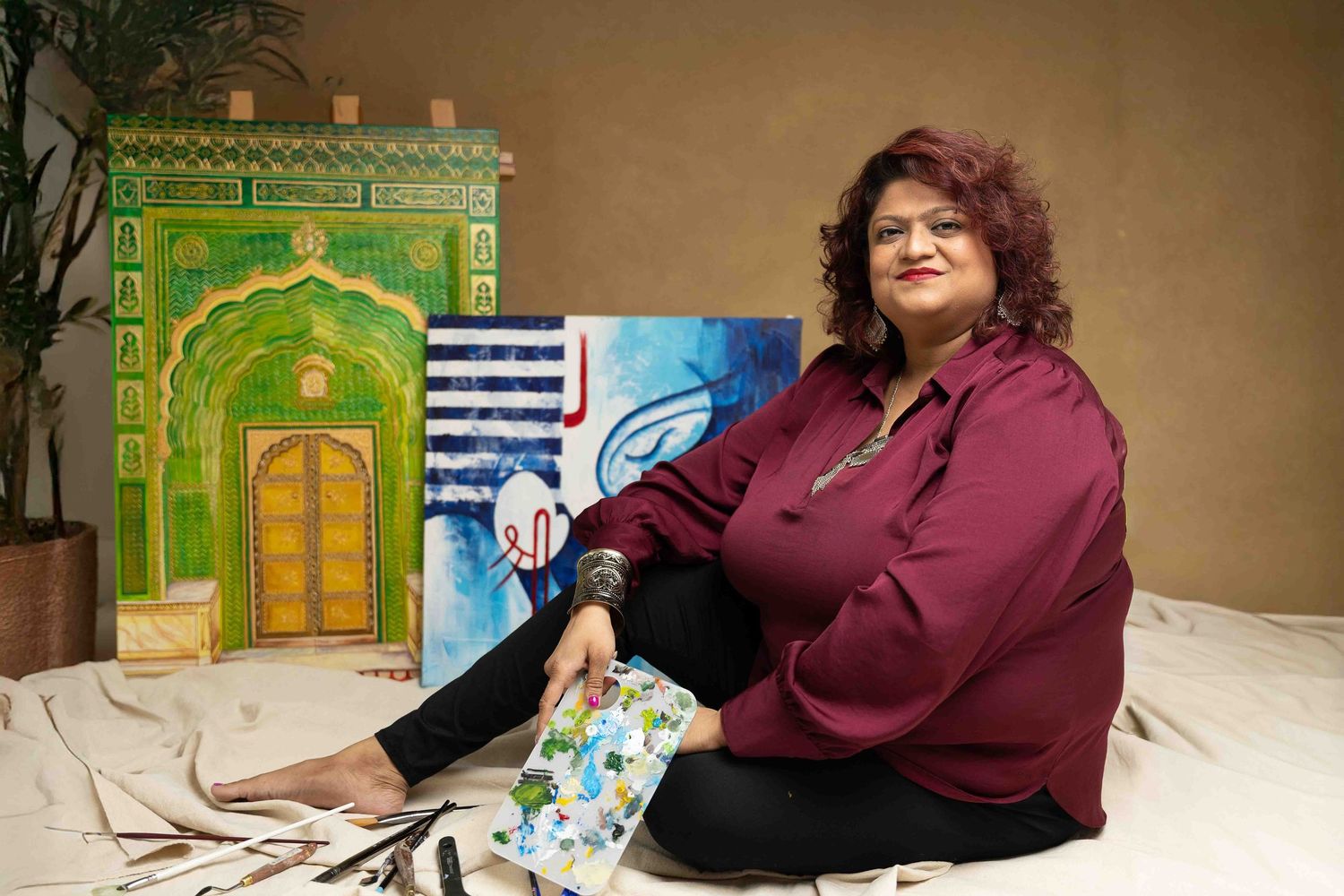Woman artist sitting beside colorful paintings including a green building and blue abstract artwork in a studio setting.