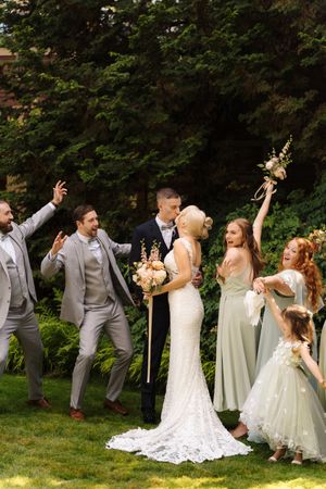 Bride and groom kissing surrounded by cheering wedding party in gray suits and sage bridesmaid dresses outdoors.