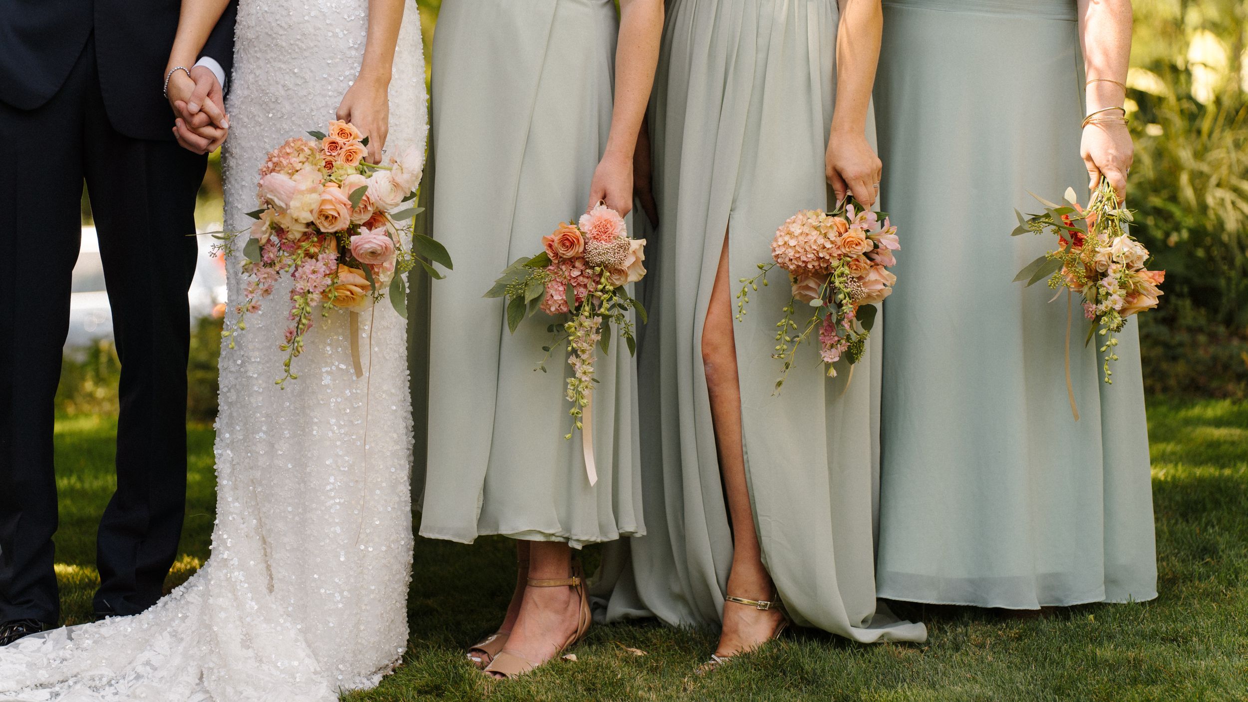 Bride in white lace gown and bridesmaids in sage green dresses holding pink floral bouquets outdoors.
