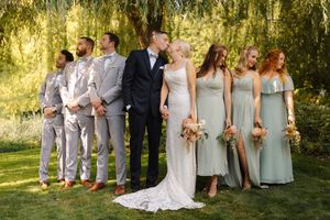 Wedding party poses outdoors under weeping willow, bride and groom kissing flanked by groomsmen in gray suits and bridesmaids in sage gowns.