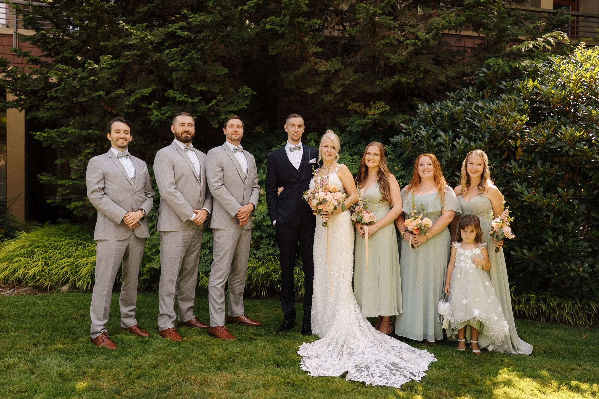 Wedding party poses outdoors, groomsmen in gray suits, bridesmaids in sage green dresses, bride and groom centered on lawn.