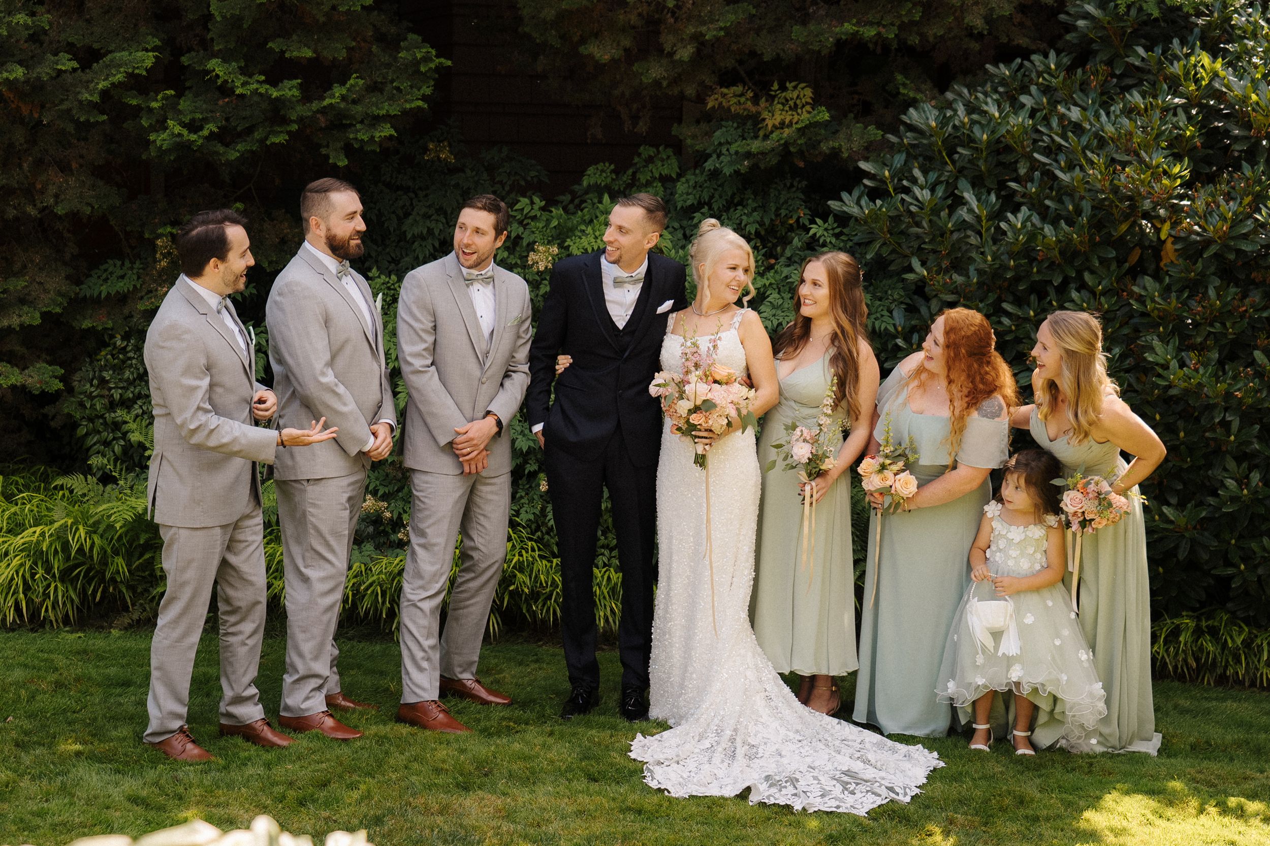 Wedding party poses outdoors on green lawn, bride and groom flanked by groomsmen in gray suits and bridesmaids in sage dresses.