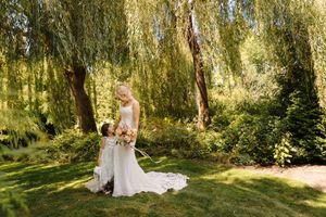 Bride in white gown holding bouquet stands under weeping willow tree in lush green garden on sunny wedding day.