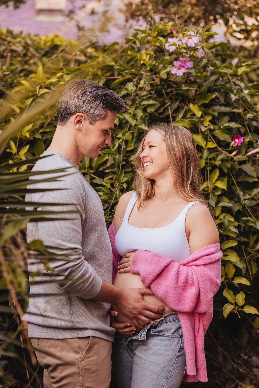 Smiling pregnant couple holding baby bump together in a lush garden with pink flowers blooming behind them.