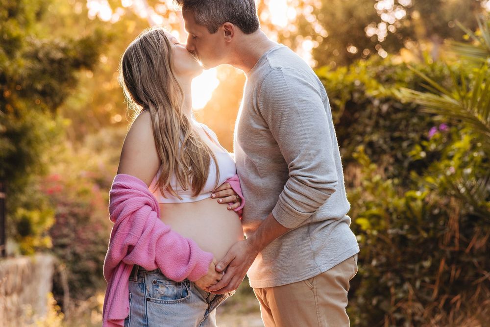 Couple kissing outdoors at golden hour, woman showing pregnant belly, surrounded by lush greenery in warm sunlight.