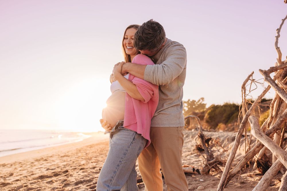 Couple embracing and laughing on a sunny beach at sunset, woman in pink top and jeans, romantic outdoor moment.