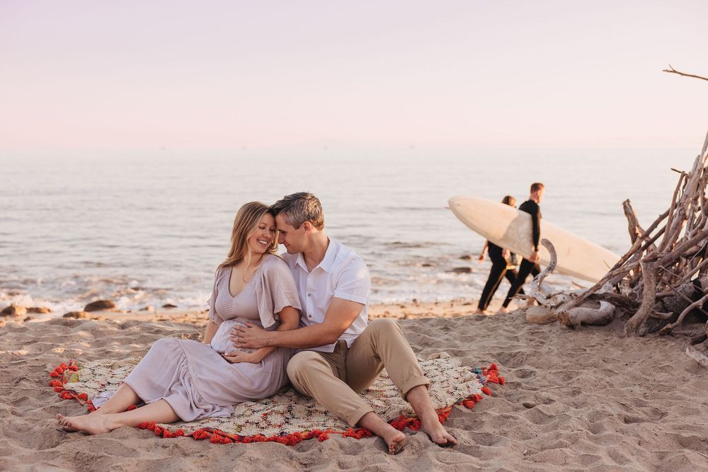 Romantic couple sitting on beach with rose petals, surfer in background at sunset.