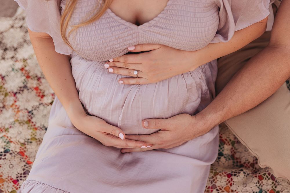 Pregnant woman in lavender dress cradling baby bump with partner's hands on a patterned blanket.