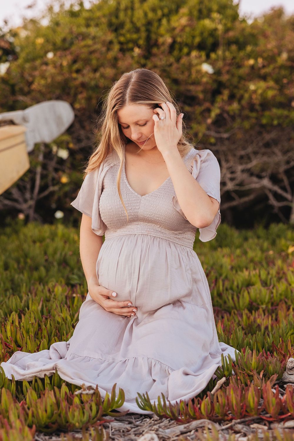 Pregnant woman in a flowing beige dress kneeling in a garden, gently cradling her baby bump during a maternity photoshoot.