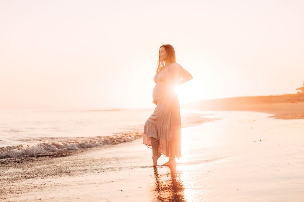 Pregnant woman in flowing dress standing on beach at golden sunset with waves gently touching the shore.