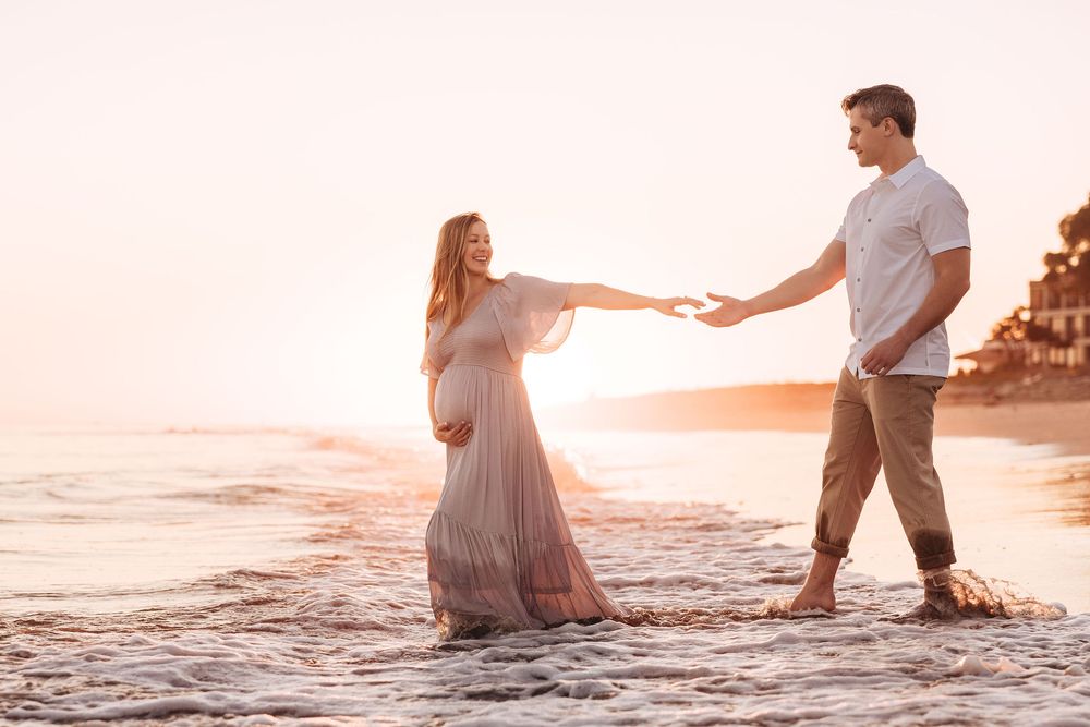 Pregnant couple holding hands on a sunlit beach at golden hour, woman in flowing pink maternity gown.