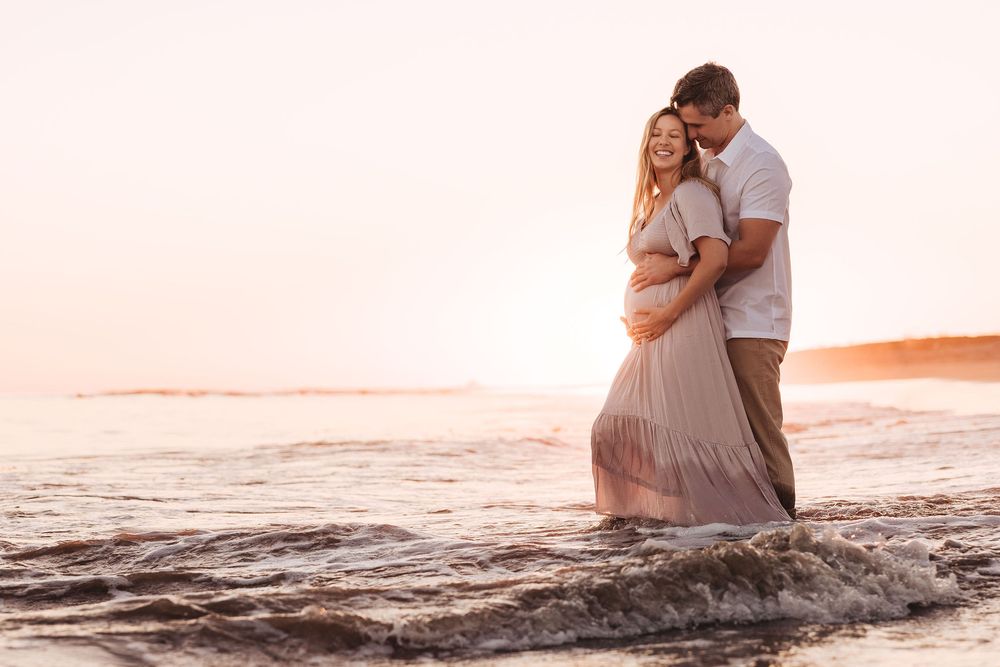 Couple embracing on beach at sunset, woman in pink maternity dress, waves washing over their feet.
