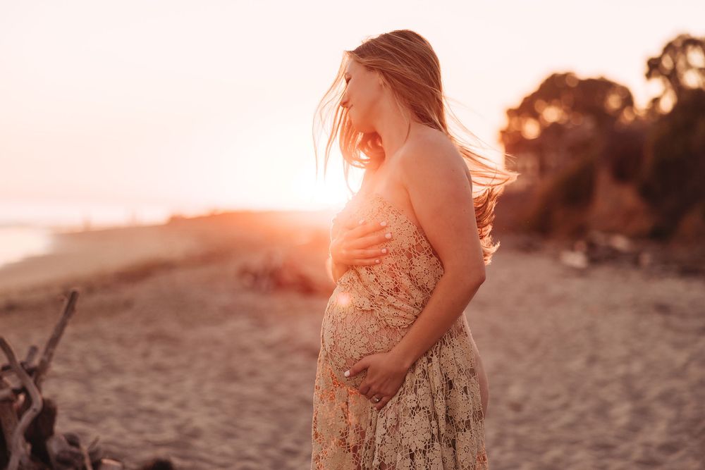 Pregnant woman in floral dress holding baby bump on beach at golden sunset with wind in her hair.