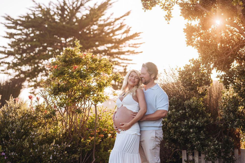 Couple embracing outdoors during golden hour, pregnant woman in white dress with partner kissing her cheek among lush green bushes.
