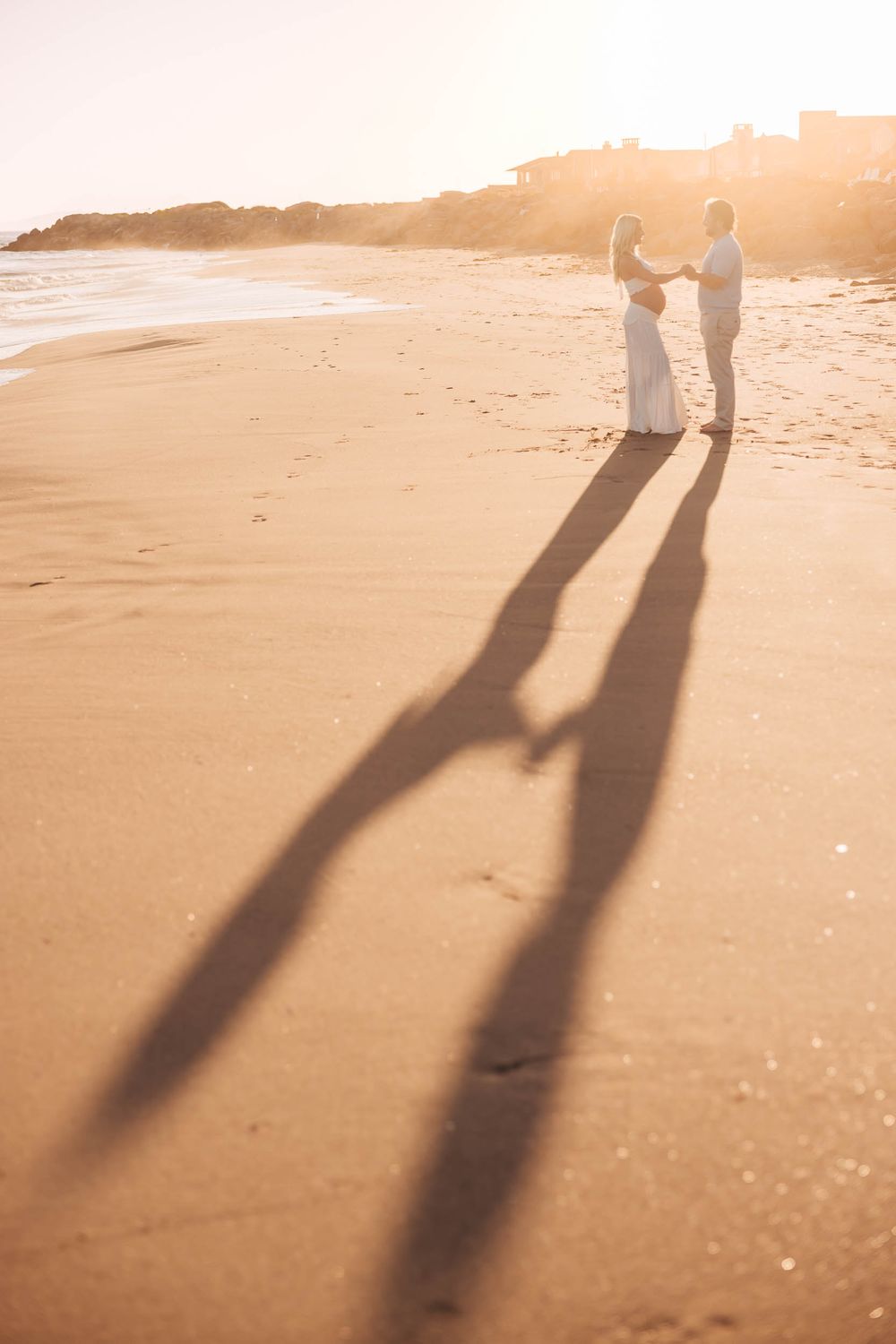 Couple in white holding hands on a sandy beach at golden sunset with long romantic shadows stretching across the shore.