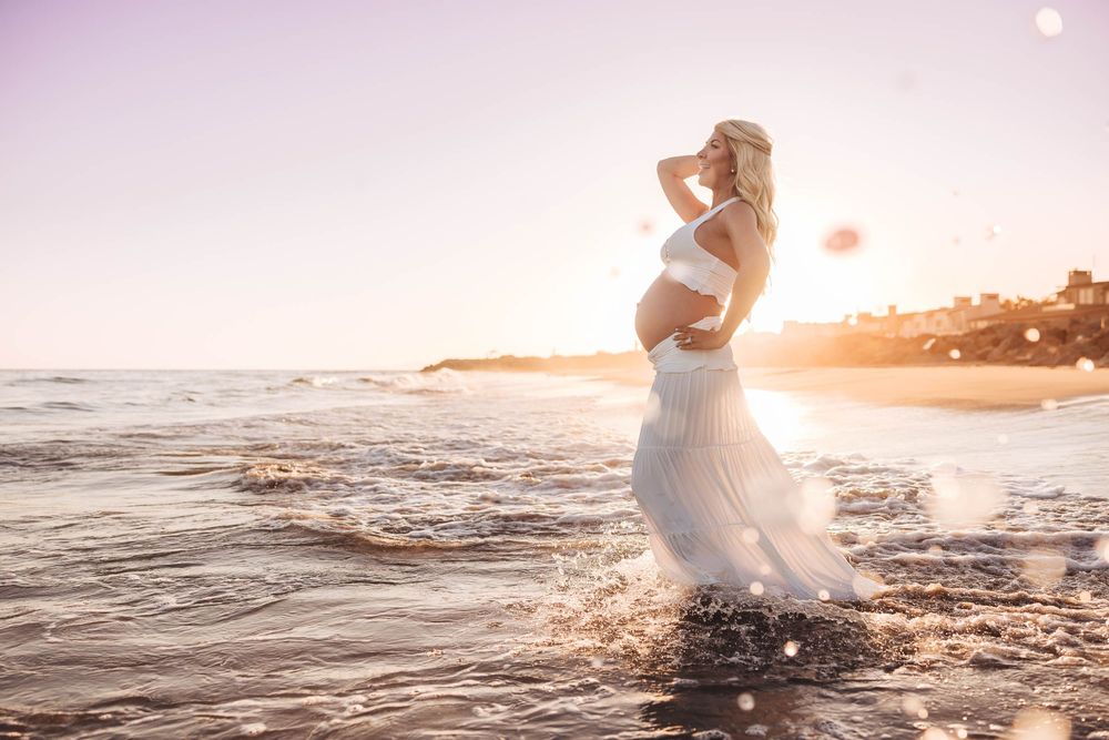 Pregnant woman in white dress standing in ocean waves at sunset on a beautiful beach.