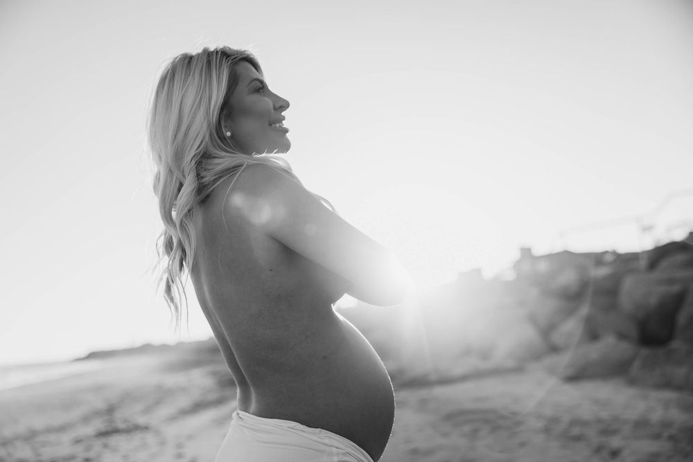Black and white photo of a pregnant woman on a beach at sunset, cradling her baby bump with sunlight glowing behind her.