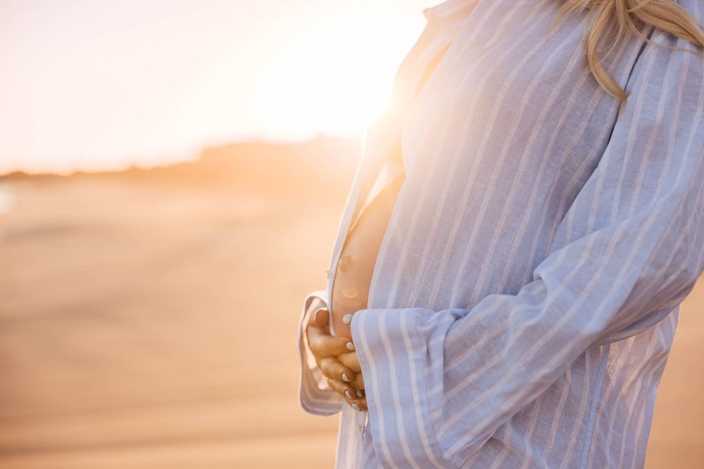 Pregnant woman in striped shirt cradling baby bump on beach at golden sunset.