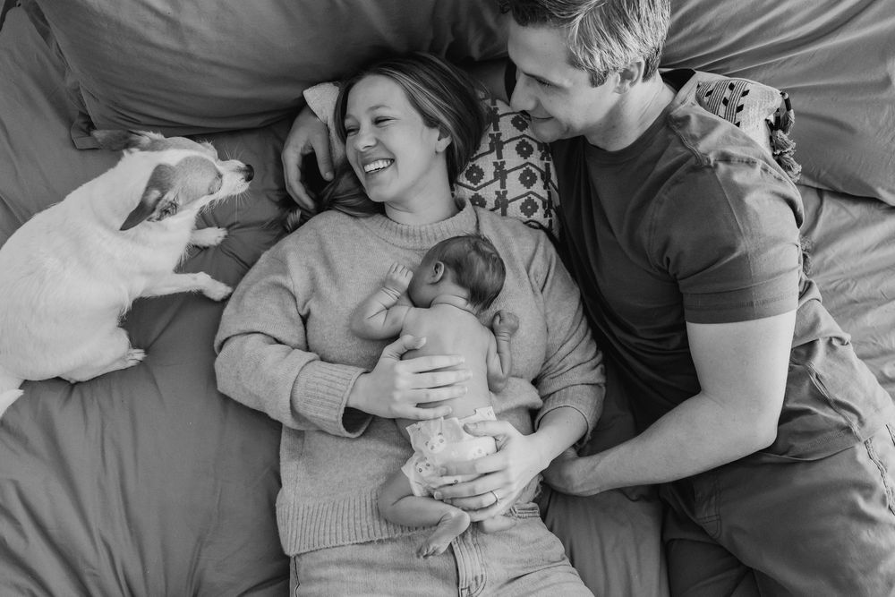 Black and white photo of smiling parents lying on bed with newborn baby and white dog in cozy home setting.