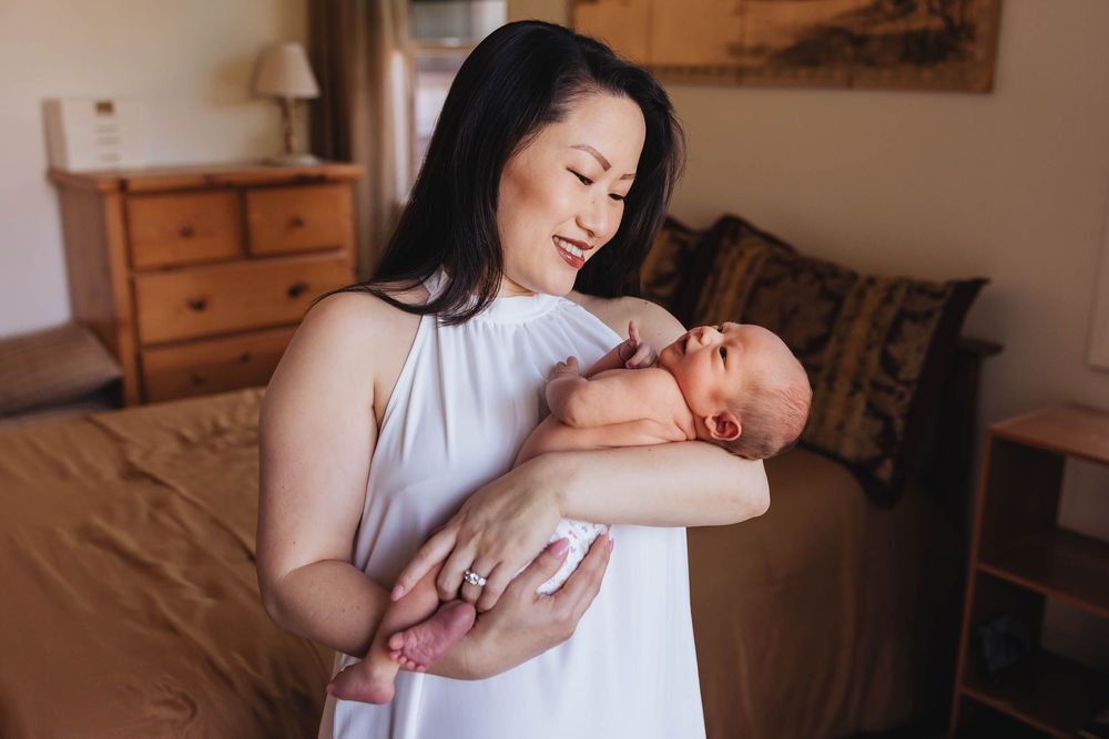 A woman in a white dress tenderly holds a newborn baby in a warmly decorated bedroom.