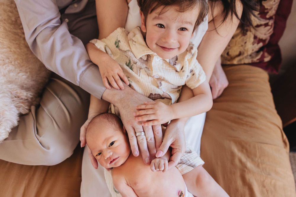 Smiling toddler holds newborn baby sibling while parents embrace them in a warm, cozy family newborn photo session.