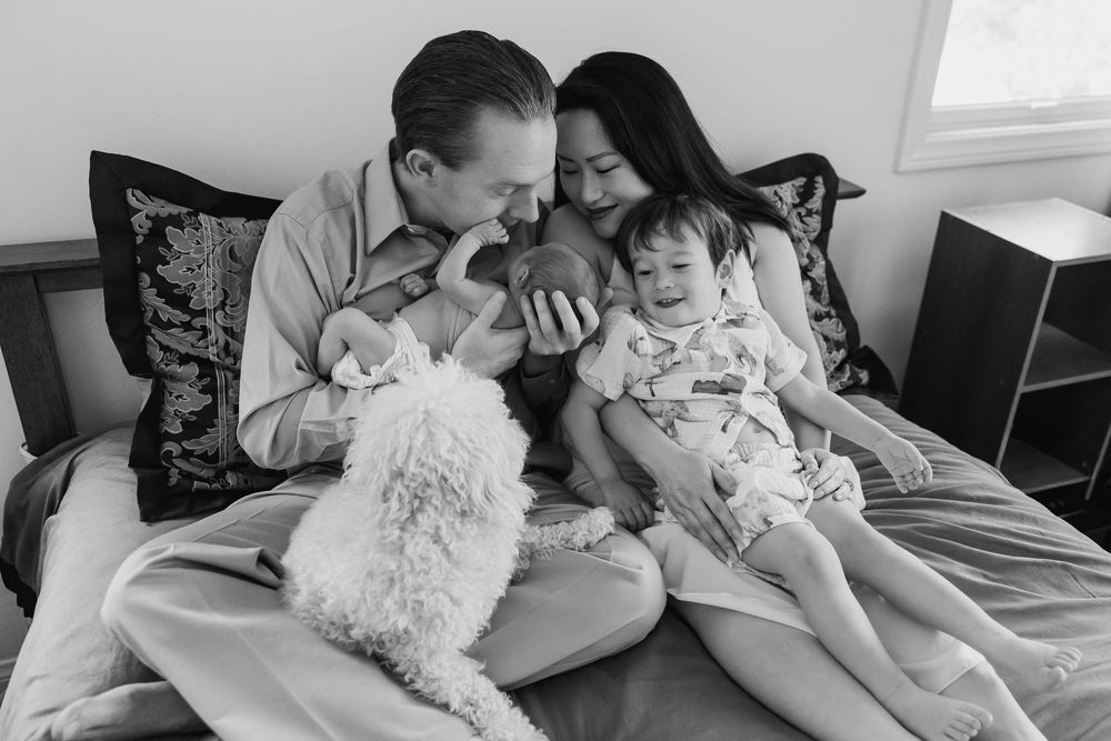 Black and white family photo of parents with newborn baby, toddler, and fluffy white dog sitting on bed.