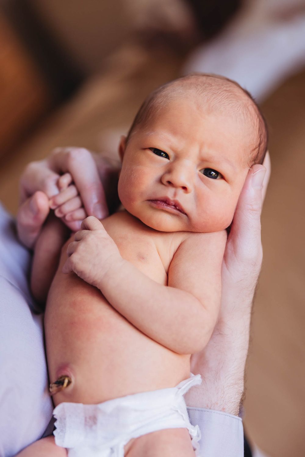 Newborn baby with furrowed brow being cradled in adult hands, wearing a white diaper.