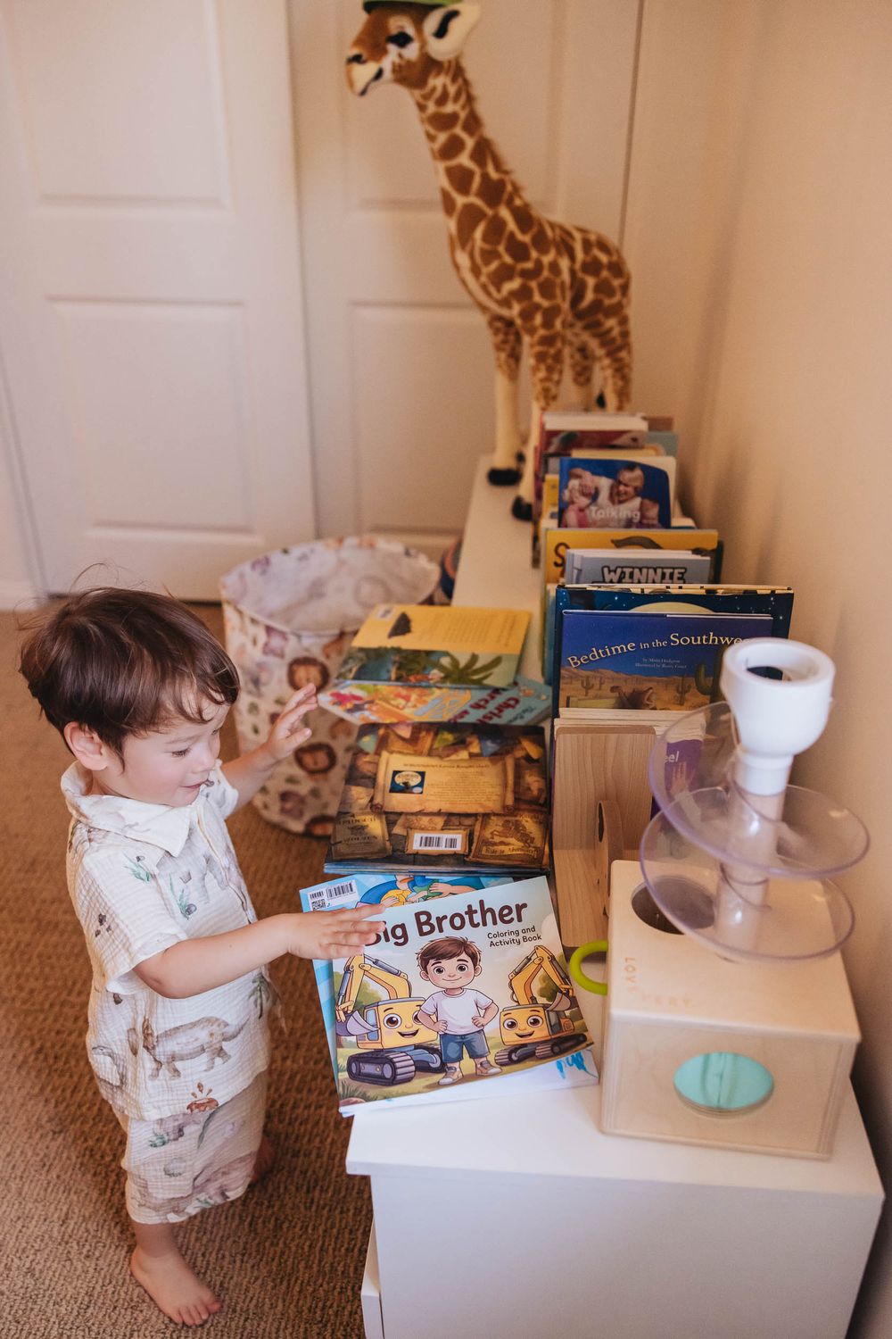 Toddler boy holding a Big Brother book beside a white dresser with children's books and a giraffe stuffed animal.