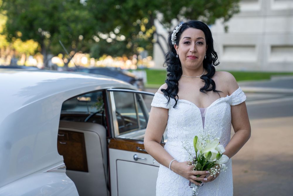 Bride in white lace off-shoulder gown holds bouquet beside vintage car outdoors.