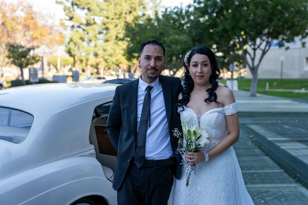 A bride in a white off-shoulder gown holding flowers stands beside a groom in a dark suit near a white limo.